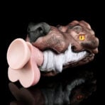 A brown dinosaur head novelty prop with a pink silicone sex toy in its mouth against a black background.