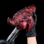 Close-up of a red, wrinkled prosthetic tongue held by gloved hands against a black background.